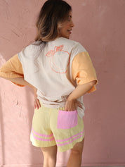Person lying on a pink surface wearing a white shirt with a red design and yellow shorts.