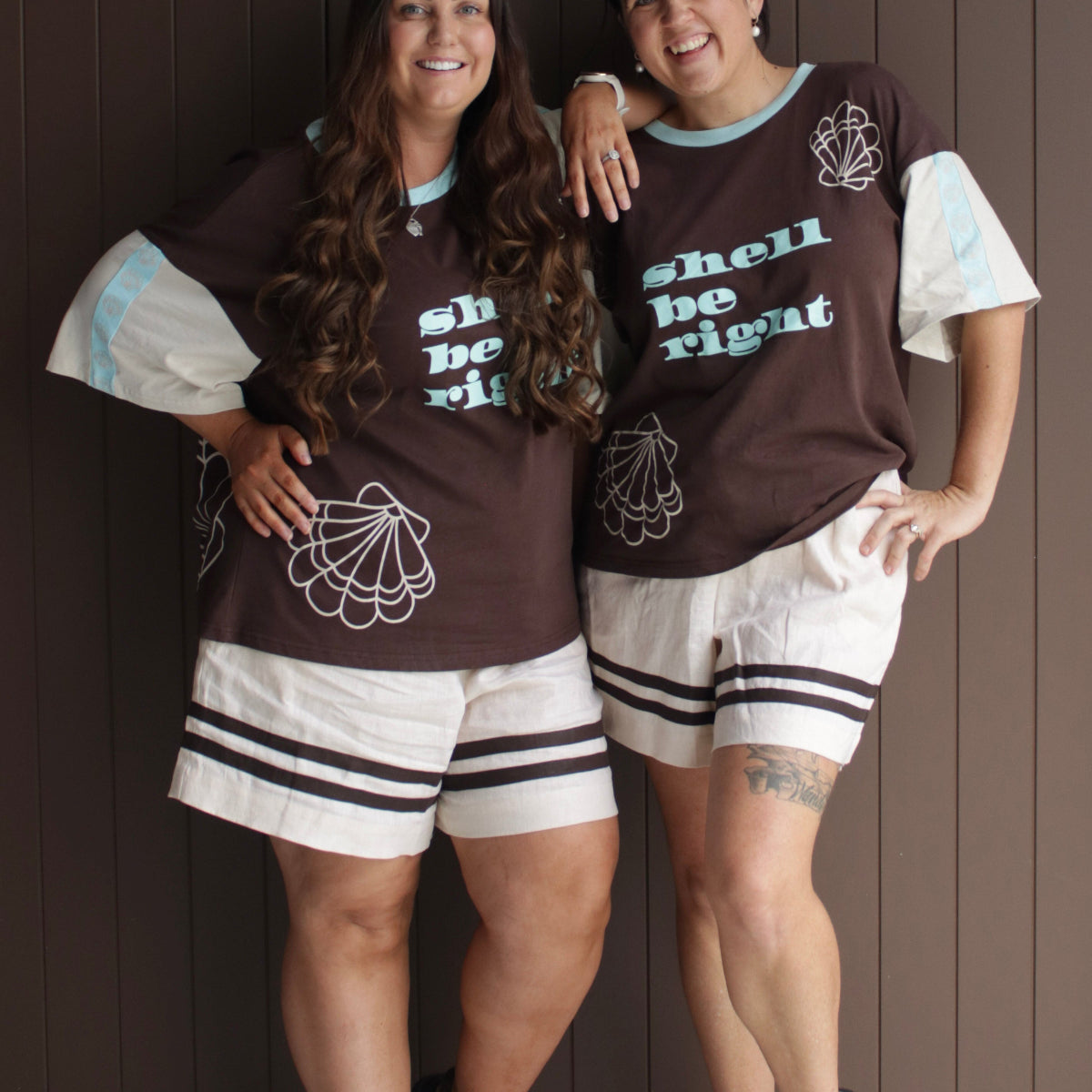 Two women wearing matching brown shirts with text and graphics, standing against a wooden panel background.