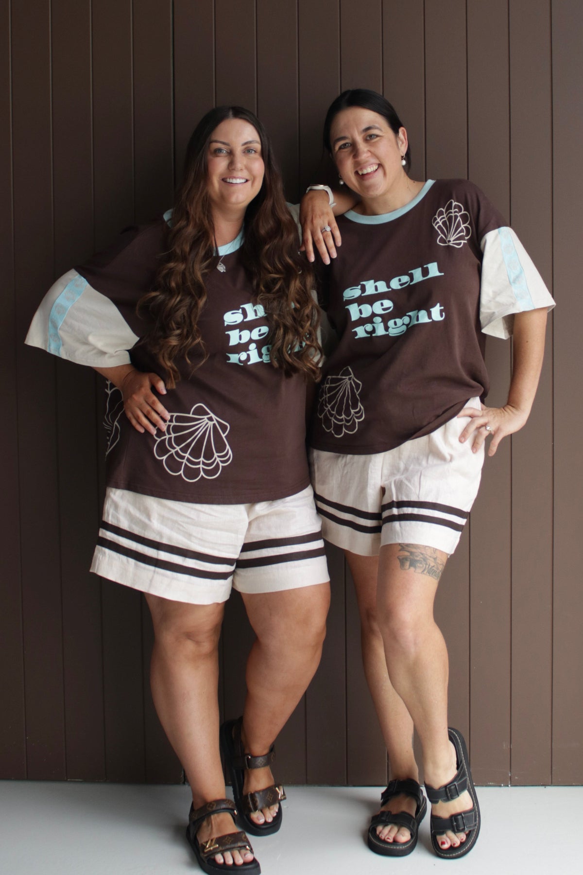 Two women wearing matching brown shirts with text and graphics, standing against a wooden panel background.