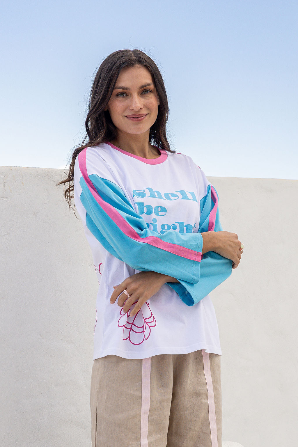 A woman wearing a white long-sleeve graphic top with bright turquoise and pink color-blocked sleeves and text reading 'shell be right', paired with beige pants, standing against a light blue sky and white wall background.