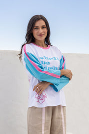 A woman wearing a white long-sleeve graphic top with bright turquoise and pink color-blocked sleeves and text reading 'shell be right', paired with beige pants, standing against a light blue sky and white wall background.