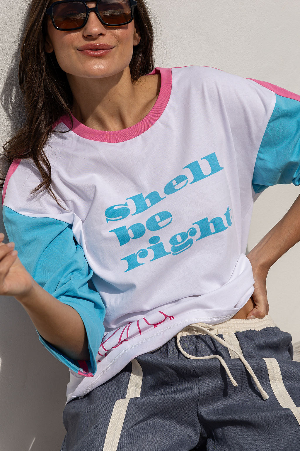 A woman wearing a white raglan t-shirt with pink and light blue color-blocked sleeves and collar, featuring the text 'shell be right' in light blue. She is also wearing dark sunglasses and gray drawstring sweatpants.