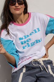 A woman wearing a white raglan t-shirt with pink and light blue color-blocked sleeves and collar, featuring the text 'shell be right' in light blue. She is also wearing dark sunglasses and gray drawstring sweatpants.
