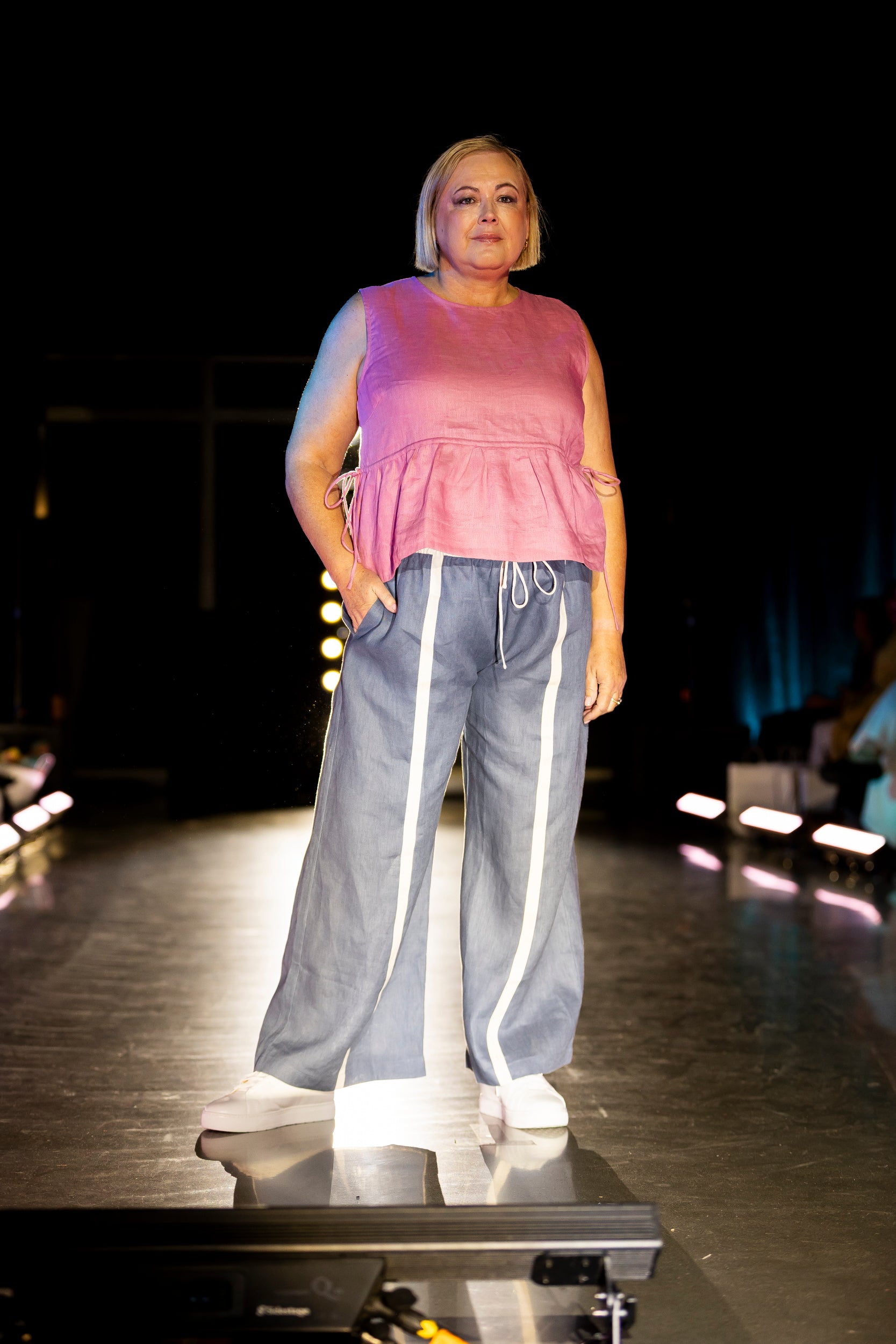 Model on a fashion runway wearing a pink sleeveless top and gray pants with white stripes.