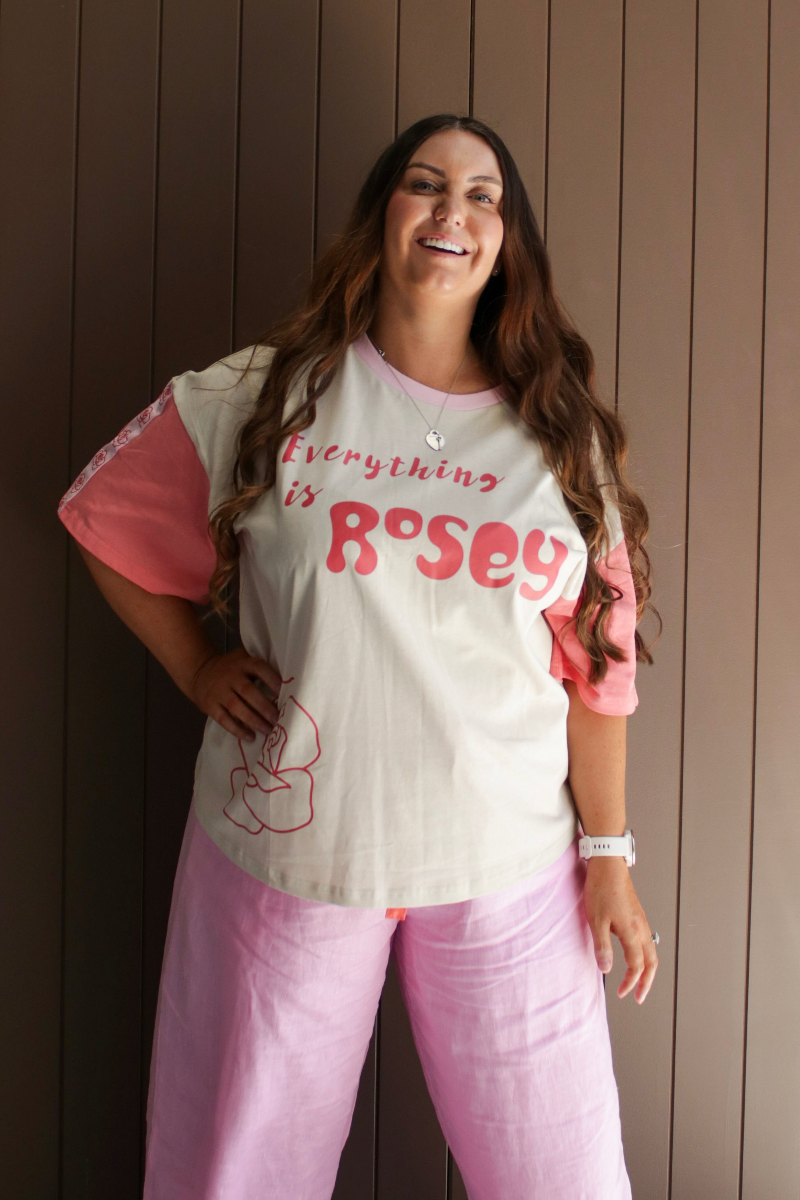 Woman wearing a 'Everything is Rosy' t-shirt and pink pants standing against a wooden wall.