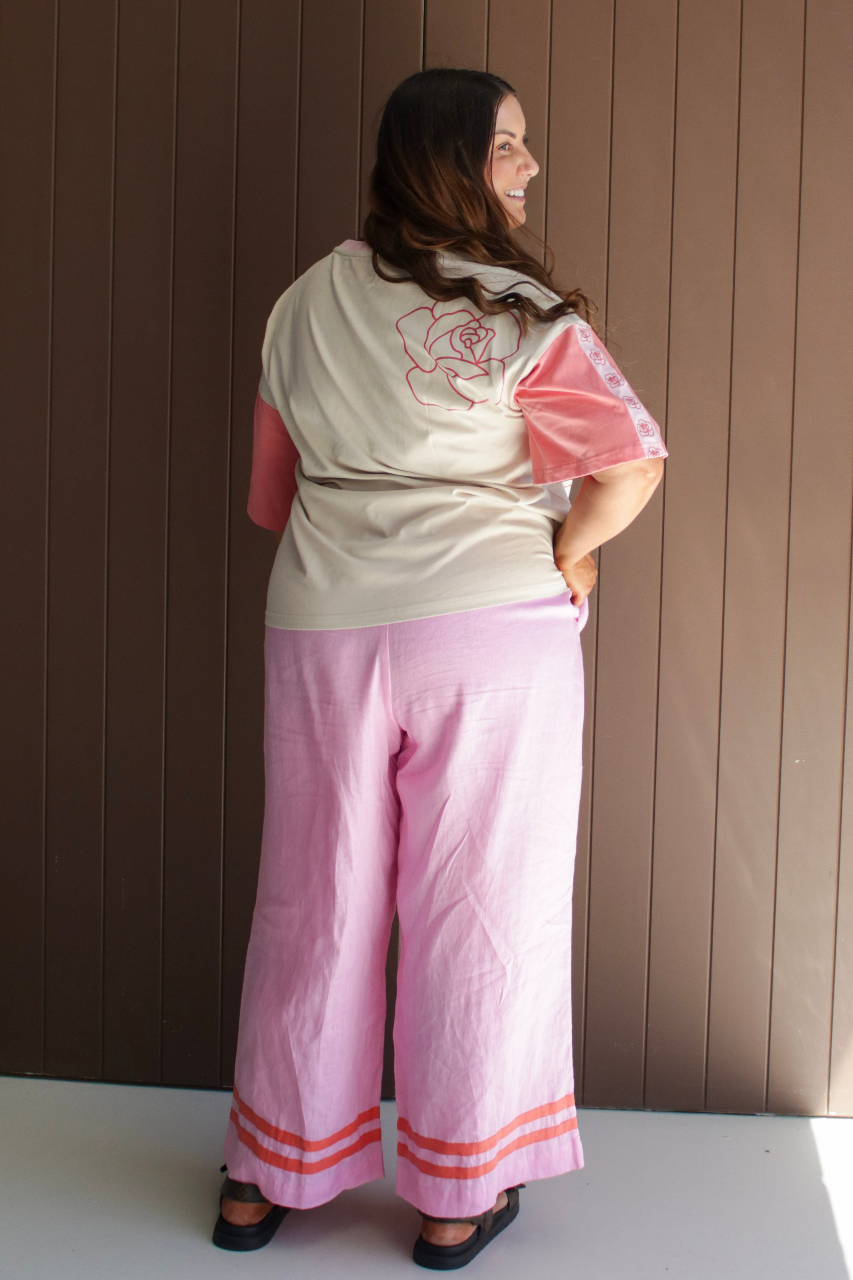 Woman wearing a light-colored top with a floral design and pink pants against a brown wall.